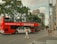 A red double-decker tour bus is driving down an urban street with a person walking across the road. The scene includes several buildings, a tall tree, and signs for tourist information and parking. The sky is partly cloudy.