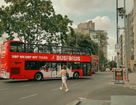 A red double-decker tour bus is driving down an urban street with a person walking across the road. The scene includes several buildings, a tall tree, and signs for tourist information and parking. The sky is partly cloudy.