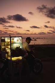 A food vendor on a motorcycle at dusk with a lit-up food display against a backdrop of a sunset sky at the beach. The sky is filled with scattered clouds, and the horizon is illuminated by the soft glow of the setting sun.
