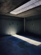 A dimly lit concrete garage with a grated window casting a beam of light onto the floor. The interior contains multiple closed doors with vertical lines and a drain on the ground.