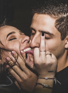 Two people are close together with one person playfully pressing their lips against the other's cheek. One person is showing their middle finger in a playful or rebellious manner. There is a close-up of their faces, both with closed eyes and smiles, suggesting a moment of friendship or affection.