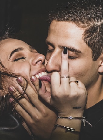 Two people are close together with one person playfully pressing their lips against the other's cheek. One person is showing their middle finger in a playful or rebellious manner. There is a close-up of their faces, both with closed eyes and smiles, suggesting a moment of friendship or affection.