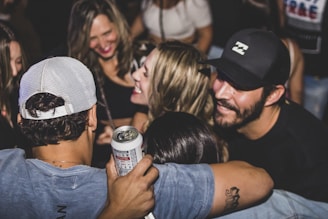 A group of people is gathered closely together, smiling and enjoying a social event. One person is holding a can, suggesting they are at a party or casual gathering. The atmosphere appears lively, with people engaging in conversation and laughter.