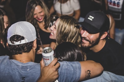 A group of people is gathered closely together, smiling and enjoying a social event. One person is holding a can, suggesting they are at a party or casual gathering. The atmosphere appears lively, with people engaging in conversation and laughter.