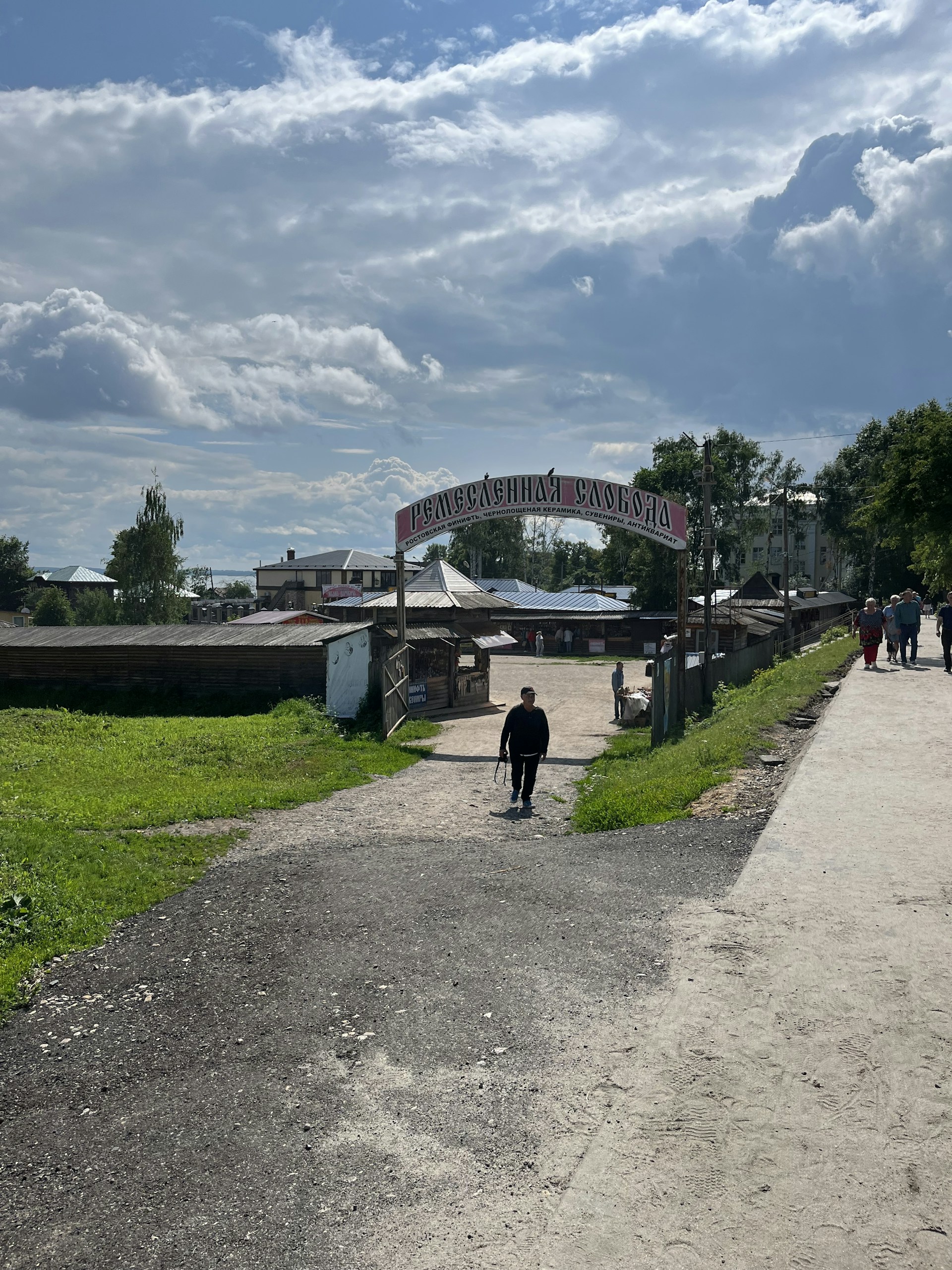 A rural scene with a pathway leading to a wooden gate labeled in Cyrillic. Beyond the gate, there are traditional style buildings. A person is walking along the path, and a few more people are visible in the background. The sky is partly cloudy, and there is a mix of greenery and paved paths.