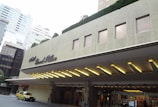 The image depicts the entrance of a building with the name 'Hotel Grand Palace' on its facade. The structure features a modern architectural design with rectangular windows and a marquee with illuminated lights. A yellow and green taxi is parked in front of the building. Surrounding skyscrapers and trimmed greenery can be seen in the background.