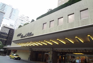 The image depicts the entrance of a building with the name 'Hotel Grand Palace' on its facade. The structure features a modern architectural design with rectangular windows and a marquee with illuminated lights. A yellow and green taxi is parked in front of the building. Surrounding skyscrapers and trimmed greenery can be seen in the background.