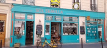 A storefront with bright turquoise blue and red accents is visible, featuring a sign that reads 'SOL SEMILLA.' The establishment appears to be a restaurant or caf&eacute; with a focus on superfoods, as indicated by the signage. Inside, people are seated at a table, and a menu board with writing is displayed outside. A yellow bicycle is parked in front of the establishment, and potted plants are placed on either side of the entrance.