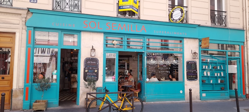 A storefront with bright turquoise blue and red accents is visible, featuring a sign that reads 'SOL SEMILLA.' The establishment appears to be a restaurant or caf&eacute; with a focus on superfoods, as indicated by the signage. Inside, people are seated at a table, and a menu board with writing is displayed outside. A yellow bicycle is parked in front of the establishment, and potted plants are placed on either side of the entrance.