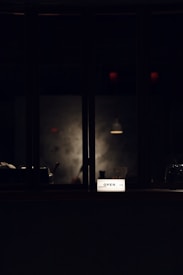 A dimly lit scene with a glowing lightbox displaying the word 'OPEN' set against a backdrop of a dark window. Faint reflections and shadows create a mysterious and inviting atmosphere.