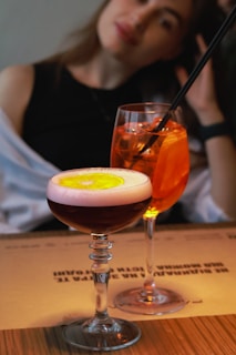 A person is seen sitting at a table with two elegant cocktails in the foreground. One of the drinks has a vibrant orange color with ice cubes and a straw, while the other is a dark-colored cocktail with a frothy top and a slice of lemon floating on it.