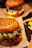 A close-up of a hamburger with a seeded bun, green and red leaf lettuce, and melted cheese. Another burger and a side of fries are blurred in the background, creating a warm and appetizing setting.