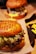 A close-up of a hamburger with a seeded bun, green and red leaf lettuce, and melted cheese. Another burger and a side of fries are blurred in the background, creating a warm and appetizing setting.