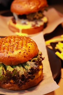 A close-up of a hamburger with a seeded bun, green and red leaf lettuce, and melted cheese. Another burger and a side of fries are blurred in the background, creating a warm and appetizing setting.