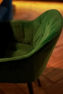 A green upholstered chair with a soft, plush texture is positioned on a wooden floor. The chair has a curved backrest and is captured in warm ambient lighting, adding a cozy atmosphere.