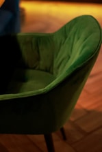 A green upholstered chair with a soft, plush texture is positioned on a wooden floor. The chair has a curved backrest and is captured in warm ambient lighting, adding a cozy atmosphere.