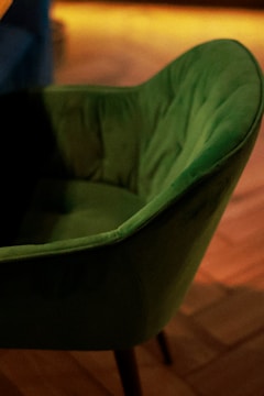 A green upholstered chair with a soft, plush texture is positioned on a wooden floor. The chair has a curved backrest and is captured in warm ambient lighting, adding a cozy atmosphere.