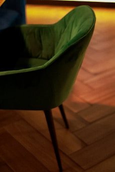 A green, upholstered chair with thin legs is positioned on a wooden parquet floor. The lighting in the room is warm, creating a cozy atmosphere with shadows and highlights on the chair.