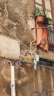 An old, weathered exterior wall in a narrow street features a faded street sign reading 'Via Isnello Sez. Tribunali'. A rusty metal balcony with potted plants and a faded cloth hanging over the ledge is visible next to a window. There are some exposed wires and a piece of cloth hanging from them.