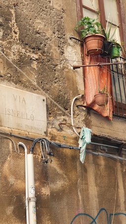 An old, weathered exterior wall in a narrow street features a faded street sign reading 'Via Isnello Sez. Tribunali'. A rusty metal balcony with potted plants and a faded cloth hanging over the ledge is visible next to a window. There are some exposed wires and a piece of cloth hanging from them.