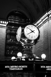 A large, ornate clock with four faces is prominently displayed atop a pedestal structure. The clock is set in a grand architectural setting with a high, intricately designed arched ceiling. Surrounding lights accentuate the metallic and classical design elements of the clock. Below, the words 'MTA Metro-North Timetables' are illuminated.