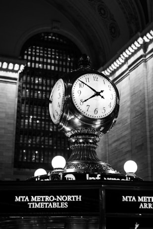A large, ornate clock with four faces is prominently displayed atop a pedestal structure. The clock is set in a grand architectural setting with a high, intricately designed arched ceiling. Surrounding lights accentuate the metallic and classical design elements of the clock. Below, the words 'MTA Metro-North Timetables' are illuminated.