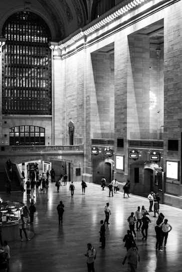 A grand hall with high ceilings and intricate architectural details, featuring strong vertical and horizontal lines from windows and arches. The hall is bustling with numerous people walking, some looking at their phones, while others are gathered in small groups. The atmosphere is lively yet somewhat serene due to the orderly movement of individuals.