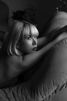 A monochrome portrait of a person with short blond hair lying on a soft, textured surface. The lighting creates a soft and intimate atmosphere, accentuating the contrast between the skin and shadows. The person's gaze is focused and slightly thoughtful.