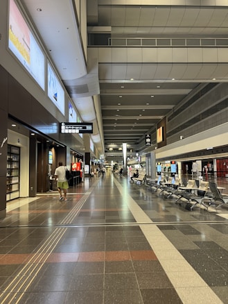 A spacious, modern airport terminal with high ceilings and overhead lighting. The floors are tiled, and there are seating areas with chairs along the sides. People can be seen walking and sitting, with some carrying bags. Signs and electronic displays are visible, providing information.