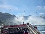 Tourists wearing bright red ponchos are gathered on a viewing platform near a large, majestic waterfall. The sky is clear with a few clouds, and several birds are flying above. The waterfall cascades down with mist rising from its base, creating a dramatic and natural scene.