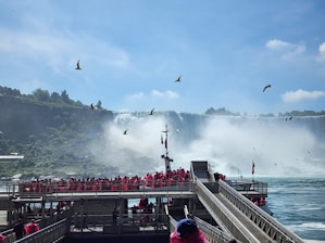 Tourists wearing bright red ponchos are gathered on a viewing platform near a large, majestic waterfall. The sky is clear with a few clouds, and several birds are flying above. The waterfall cascades down with mist rising from its base, creating a dramatic and natural scene.