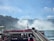 Tourists wearing bright red ponchos are gathered on a viewing platform near a large, majestic waterfall. The sky is clear with a few clouds, and several birds are flying above. The waterfall cascades down with mist rising from its base, creating a dramatic and natural scene.