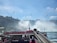 Tourists wearing bright red ponchos are gathered on a viewing platform near a large, majestic waterfall. The sky is clear with a few clouds, and several birds are flying above. The waterfall cascades down with mist rising from its base, creating a dramatic and natural scene.