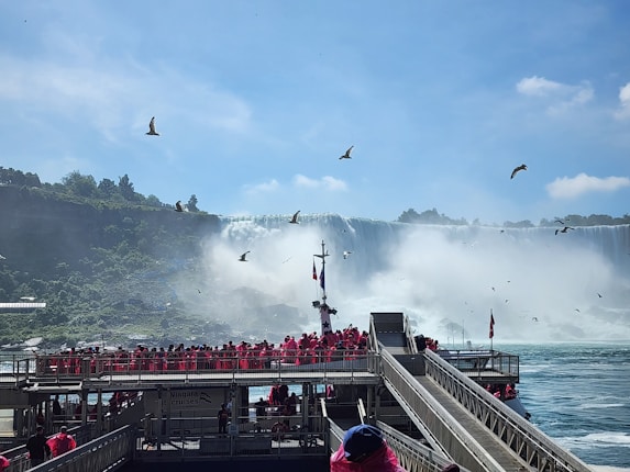 Tourists wearing bright red ponchos are gathered on a viewing platform near a large, majestic waterfall. The sky is clear with a few clouds, and several birds are flying above. The waterfall cascades down with mist rising from its base, creating a dramatic and natural scene.