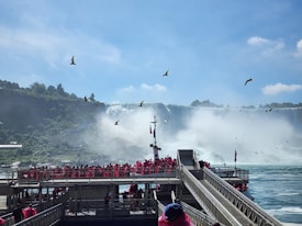 Tourists wearing bright red ponchos are gathered on a viewing platform near a large, majestic waterfall. The sky is clear with a few clouds, and several birds are flying above. The waterfall cascades down with mist rising from its base, creating a dramatic and natural scene.