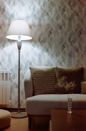 A cozy living room with a tall floor lamp emitting soft light, standing beside a comfortable sofa adorned with patterned cushions. The background features a geometric textured wallpaper, and there is a small table with a glass containing delicate flowers.