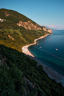 A coastal landscape featuring a lush, green hillside that slopes down to a secluded beach lined with sun loungers. The clear blue sea stretches into the distance, meeting a bright, cloudless sky. Nearby boats can be seen anchored in the water.