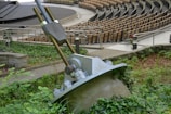 A large metal cable anchor embedded in a concrete base is surrounded by greenery. In the background, an empty amphitheater with multiple rows of wooden seating is visible. The scene is outdoors, with pathways and metal railings.