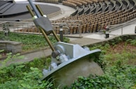 A large metal cable anchor embedded in a concrete base is surrounded by greenery. In the background, an empty amphitheater with multiple rows of wooden seating is visible. The scene is outdoors, with pathways and metal railings.