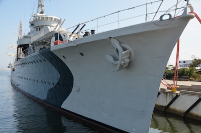 A large naval ship is docked beside a concrete pier. The ship is painted grey with darker accents and features a prominent anchor on its side. Several structures and masts are visible on top of the ship. The water is calm, and there are ropes tying the ship to the dock. In the background, parts of a building and some greenery are visible.