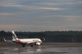 A commercial airplane with the Jetstar logo is taxiing on a wet runway at an airport. The aircraft is positioned against a backdrop of a cloudy sky and distant trees.