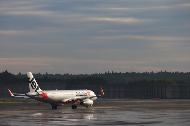 A commercial airplane with the Jetstar logo is taxiing on a wet runway at an airport. The aircraft is positioned against a backdrop of a cloudy sky and distant trees.