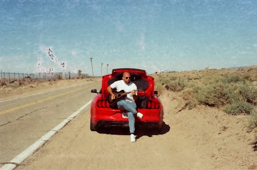 A person is sitting on a red car with an open trunk, playing a guitar on the side of a deserted road. The landscape is arid with sparse vegetation and a clear blue sky.