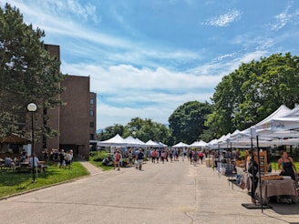 A street market scene with several white tents set up along a pathway, surrounded by lush green trees and a brick building on the left. People are walking around and browsing the stalls, some of which appear to be selling crafts or food items. The sky is clear with a few scattered clouds, suggesting a sunny day.