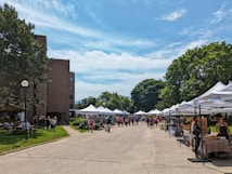 A street market scene with several white tents set up along a pathway, surrounded by lush green trees and a brick building on the left. People are walking around and browsing the stalls, some of which appear to be selling crafts or food items. The sky is clear with a few scattered clouds, suggesting a sunny day.