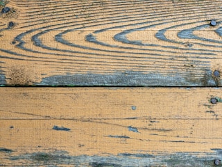 A close-up view of weathered wooden planks with visible grain patterns and several dark screws embedded at intervals. The surface shows signs of aging, with some areas appearing worn and slightly discolored. The wood displays a mix of brown and grey tones with patches of green moss or mold in some spots.