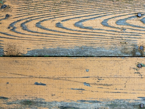 A close-up view of weathered wooden planks with visible grain patterns and several dark screws embedded at intervals. The surface shows signs of aging, with some areas appearing worn and slightly discolored. The wood displays a mix of brown and grey tones with patches of green moss or mold in some spots.