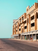 A long, empty road stretches beside a large building with ornate architecture, featuring archways and geometric designs. The building, which is under construction or renovation, is illuminated by bright sunlight against a clear, turquoise sky. Scaffolding and construction materials are visible, suggesting ongoing work.