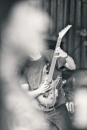 A person playing an electric guitar with a detailed wood finish, partially obscured by what appears to be smoke or a blurred element in the foreground. The image has a high contrast and a monochromatic tone, emphasizing textures and shadows.