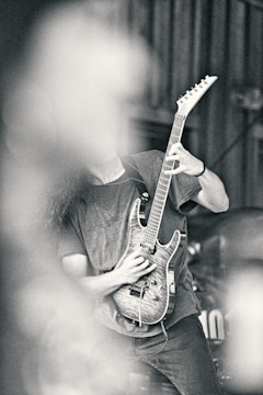 A person playing an electric guitar with a detailed wood finish, partially obscured by what appears to be smoke or a blurred element in the foreground. The image has a high contrast and a monochromatic tone, emphasizing textures and shadows.